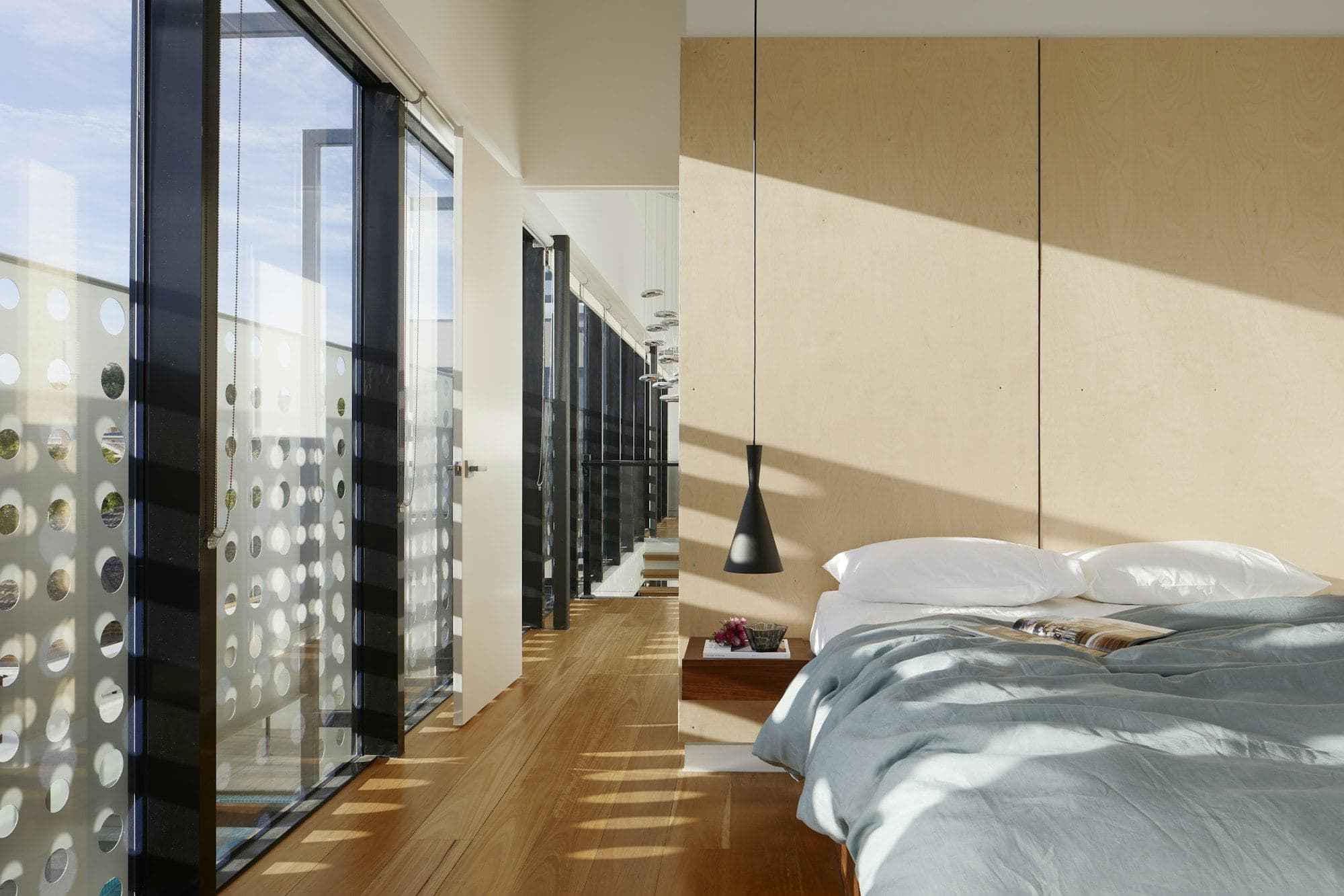 Bedroom in afternoon sun with timber floor, pale plywood wall and a perforated screen outside the glazing