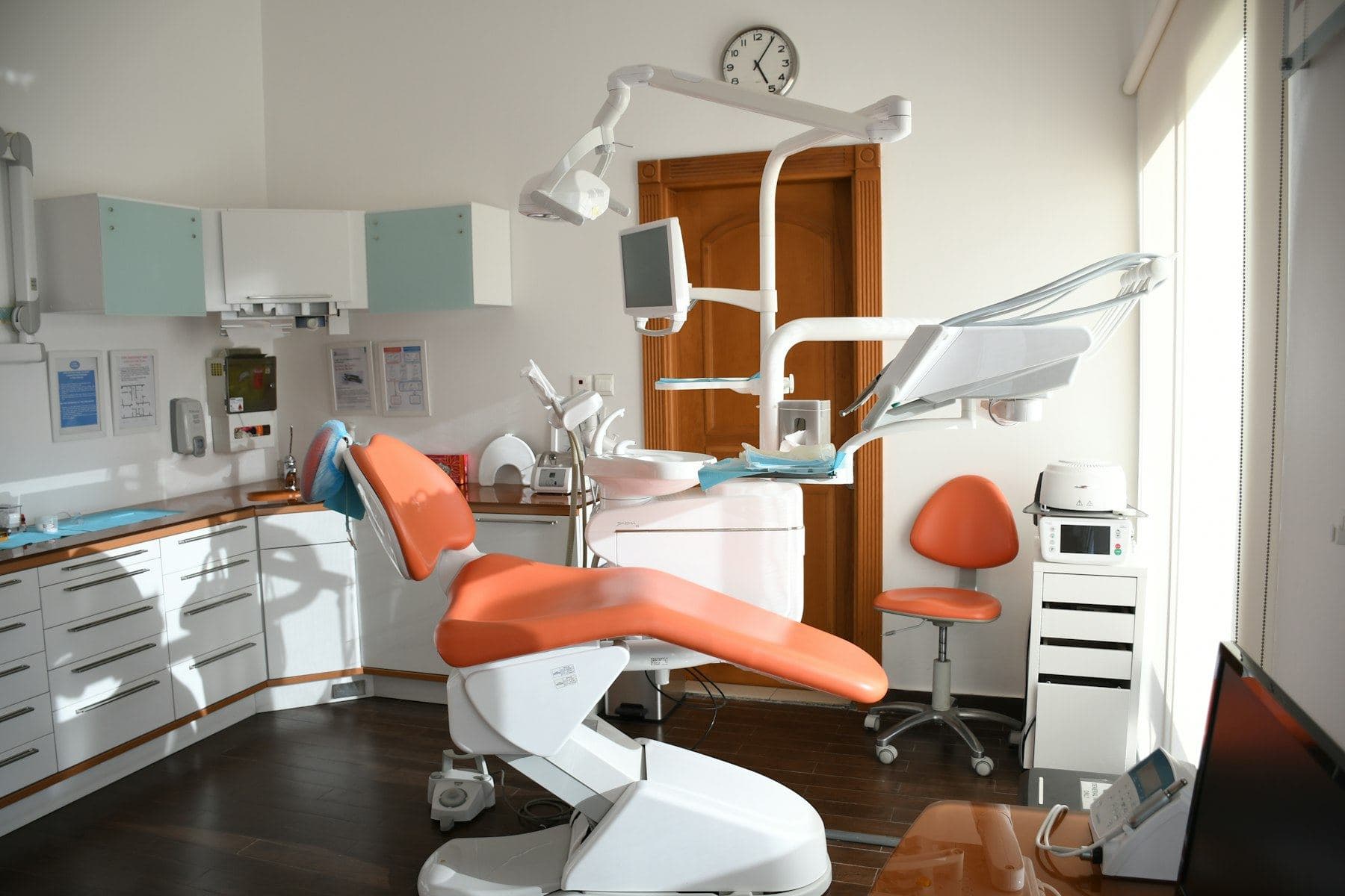 A bright treatment room with an orange dental chair, white cabinets and a large window