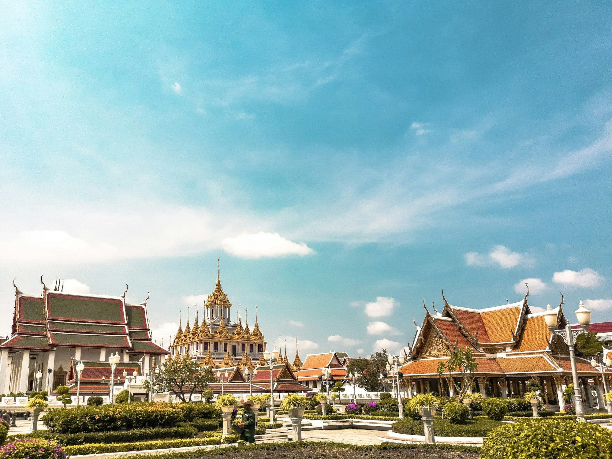 โลหะปราสาท วัดราชนัดดาราม ใต้ท้องฟ้าสีฟ้า ด้านหน้าเป็นสวนและอาคารทรงไทย