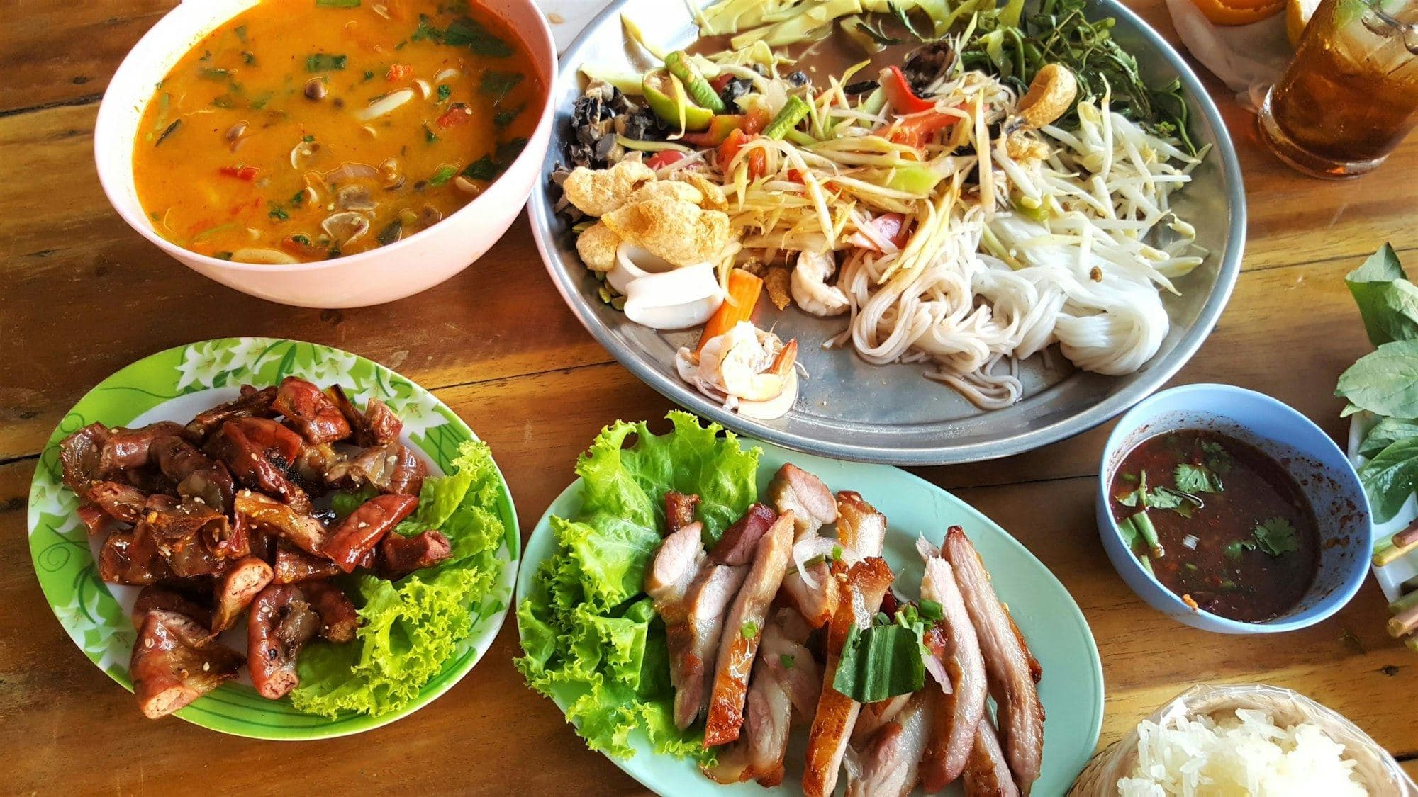 อาหารไทยหลายจานบนโต๊ะไม้ ต้มยำ ยำ หมูย่าง และผักสด