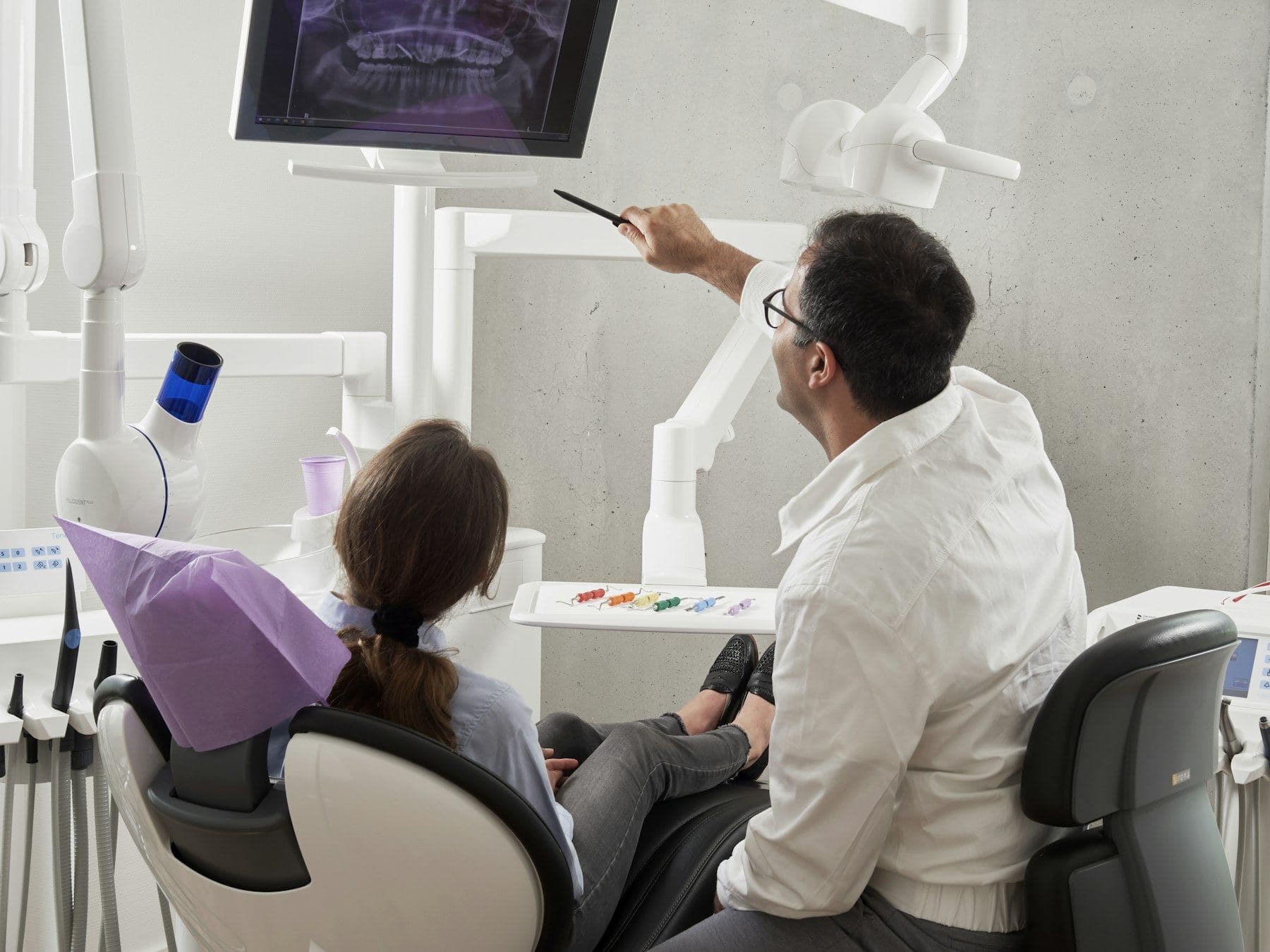 A dentist pointing at a dental X-ray on a screen for a patient sitting in the chair